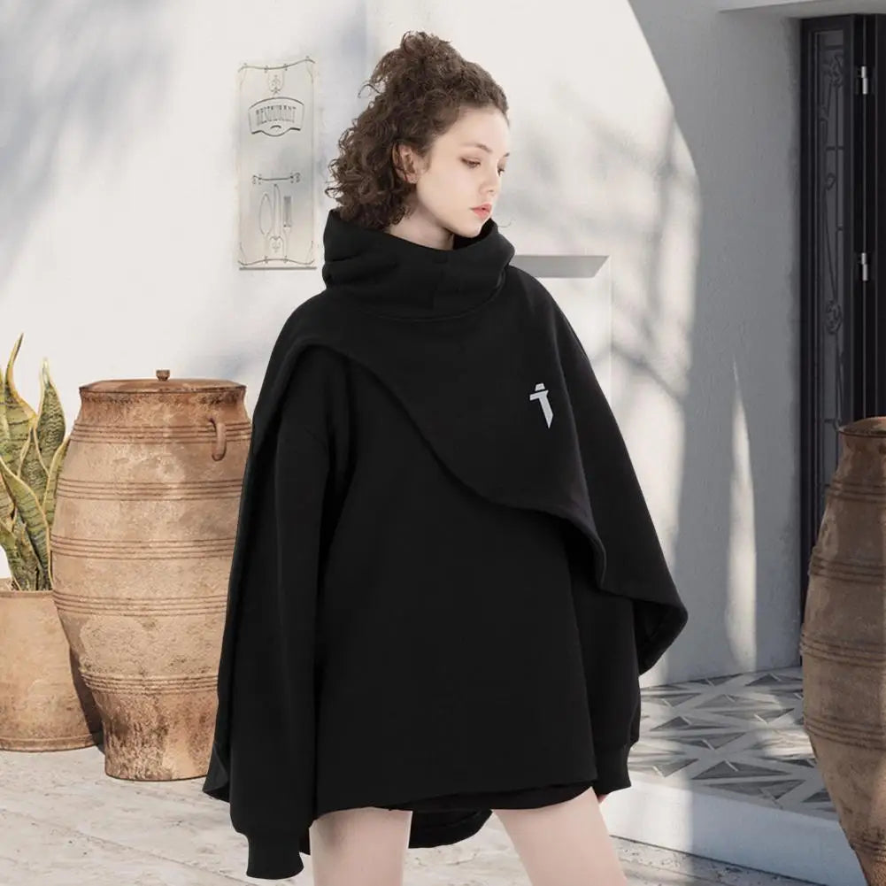 Woman wearing a black poncho with a white logo in an outdoor setting with large pots and a light background.